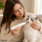 A woman sitting on a sofa gently using the white Petifo Detixy dental cleaner on the teeth of a relaxed, fluffy white and grey cat.